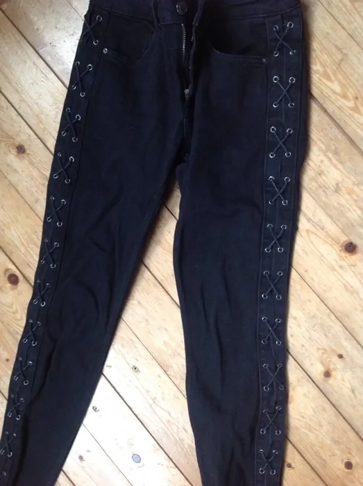 Schmale schwarze Jeans mit seitlicher Schnürung und Ösen, ausgebreitet auf einem Holzboden